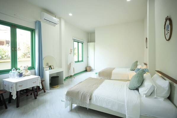 A hotel room with two beds, a desk with a chair, a window with curtains, an air conditioning unit, and a small table with a teddy bear.