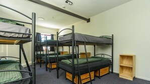 A dormitory room with bunk beds, a small wooden shelf, and a window. - The Rotten Apple Back Packers (Hastings)