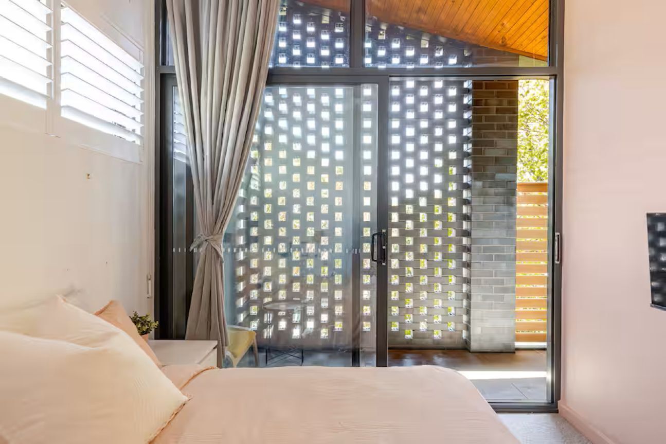 Een slaapkamer met een bed, een raam met zonwering, een gordijn en een schuifdeur van glas die uitkomt op een buitenruimte.