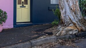 Un bâtiment avec une porte jaune, un grand arbre devant et un mur rose sur le côté gauche.