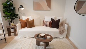 Un salon moderne avec un canapé blanc, de nombreux coussins, une petite table d’appoint en bois, une lampe sur pied et une grande plante en pot.