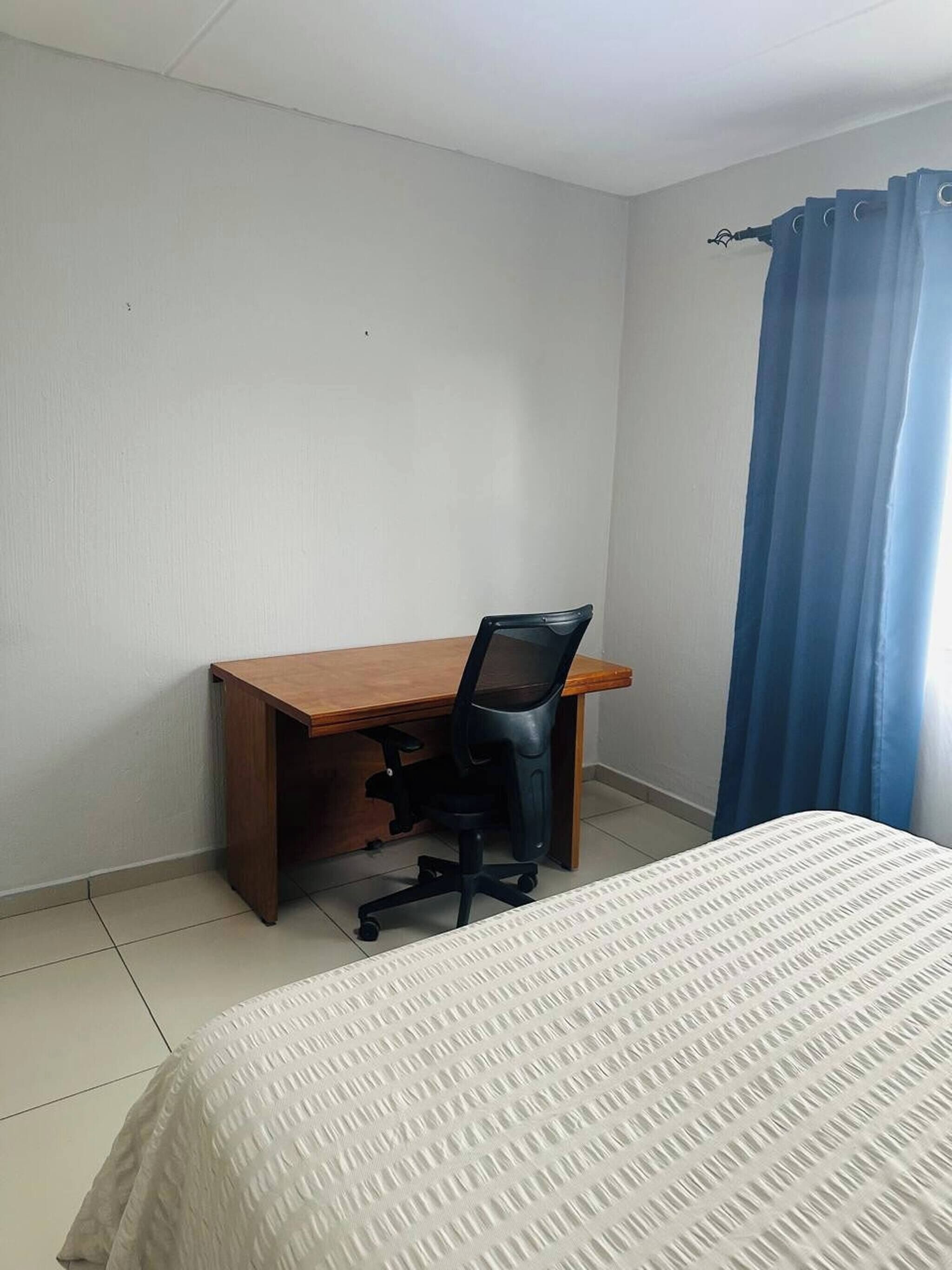 A room with a bed, a wooden desk, a black office chair, and a blue curtain.