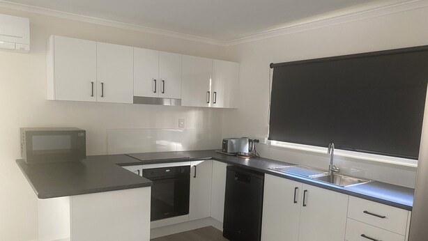 A modern kitchen with white cabinets, a black countertop, a dishwasher, a coffee maker, and a window with a black blind. - Spacious modern Beachside 2 Bedroom Unit. (Pialba)