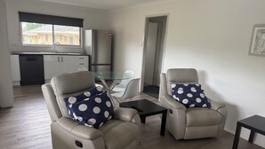A living room with two reclining chairs, a glass coffee table, and a kitchen in the background. - Spacious modern Beachside 2 Bedroom Unit. (Pialba)
