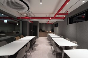 A modern conference room with white tables and chairs, a kitchen area, and red pipes on the ceiling. - OstelloMaree (Seoul)