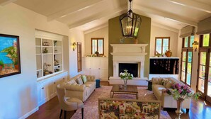 A spacious living room with a fireplace, a large painting, and a view of greenery through the windows.