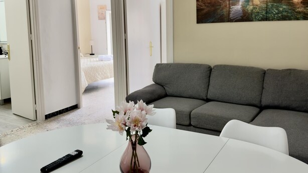 Un salón con un sofá gris, una mesa blanca con un jarrón de flores y un cuadro de una cueva y agua en la pared.