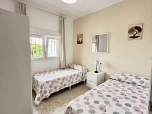 Ein Schlafzimmer mit zwei Einzelbetten, einem Nachttisch mit Lampe, einem Fenster mit Vorhängen und Wandbildern.