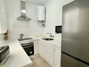 Une cuisine compacte dotée de meubles blancs, d’un réfrigérateur en acier inoxydable, d’un four encastré, d’un four à micro-ondes, d’un évier et d’un chauffe-eau.