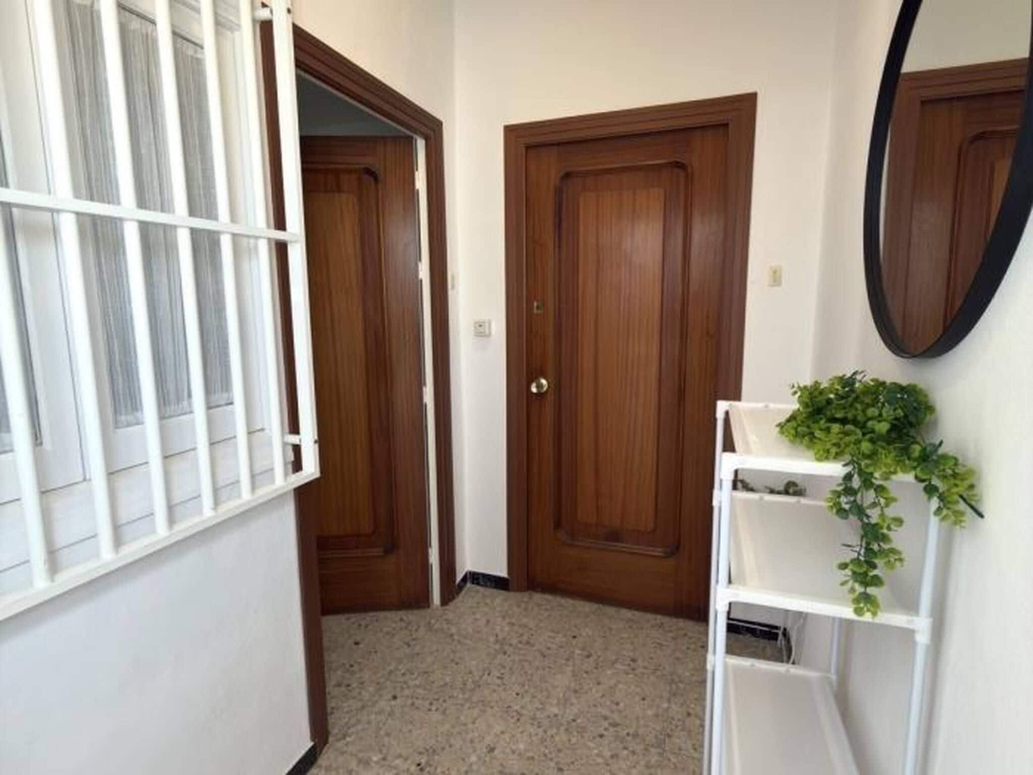Un couloir avec deux portes en bois, une étagère blanche ornée d’une plante et une fenêtre avec des stores blancs.