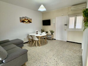 Ein kompakter Wohnraum mit einer Couch, einem Esstisch mit Stühlen, einem an der Wand montierten Fernseher und einer Glastür.