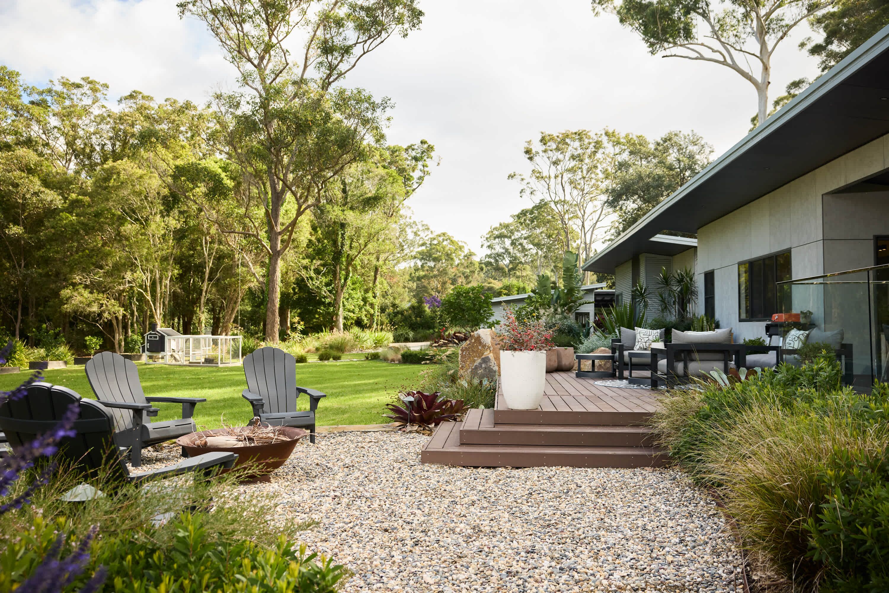 A modern outdoor patio with Adirondack chairs, a fire pit, and a deck leading to a house with large windows.