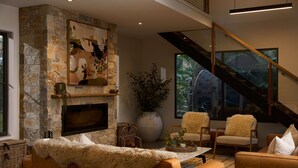 A modern living room with a stone fireplace, a flat-screen TV, comfortable seating, and a staircase with glass railings. - The Pavilion, Wamberal (Wamberal)