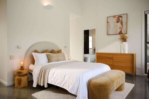 A modern bedroom with a curved bed, bedside lamps, a wooden nightstand, a bench, a dresser, and a painting on the wall.
