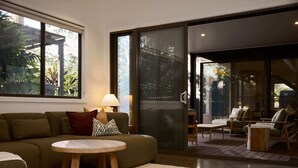 A modern living room with a sofa, round table, and chairs, large windows, and a sliding glass door leading to an outdoor area. - The Pavilion, Wamberal (Wamberal)