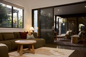 A modern living room with a sofa, round table, and chairs, large windows, and a sliding glass door leading to an outdoor area.