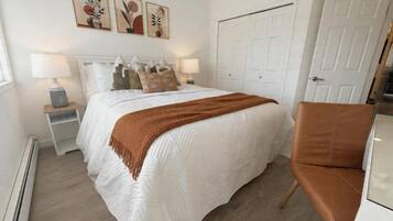 Une chambre à coucher comprenant un lit, deux tables de chevet, un fauteuil marron, une porte blanche et trois œuvres d’art accrochées au mur.
