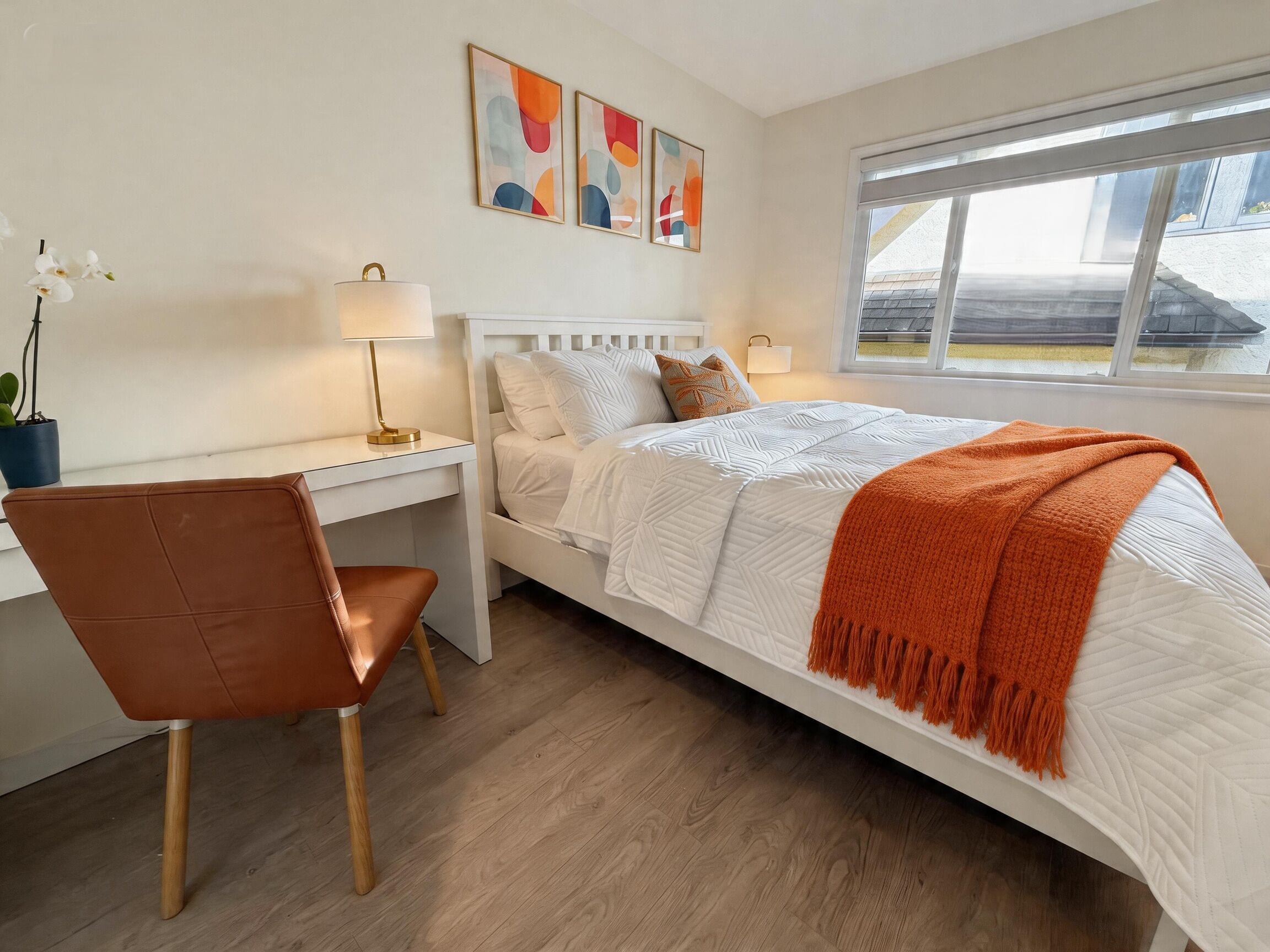 A bedroom with a bed, desk, chair, lamp, artwork, and a window.