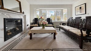 A room with a fireplace, a dining table with chairs, a sofa, and a large window with a view of trees.