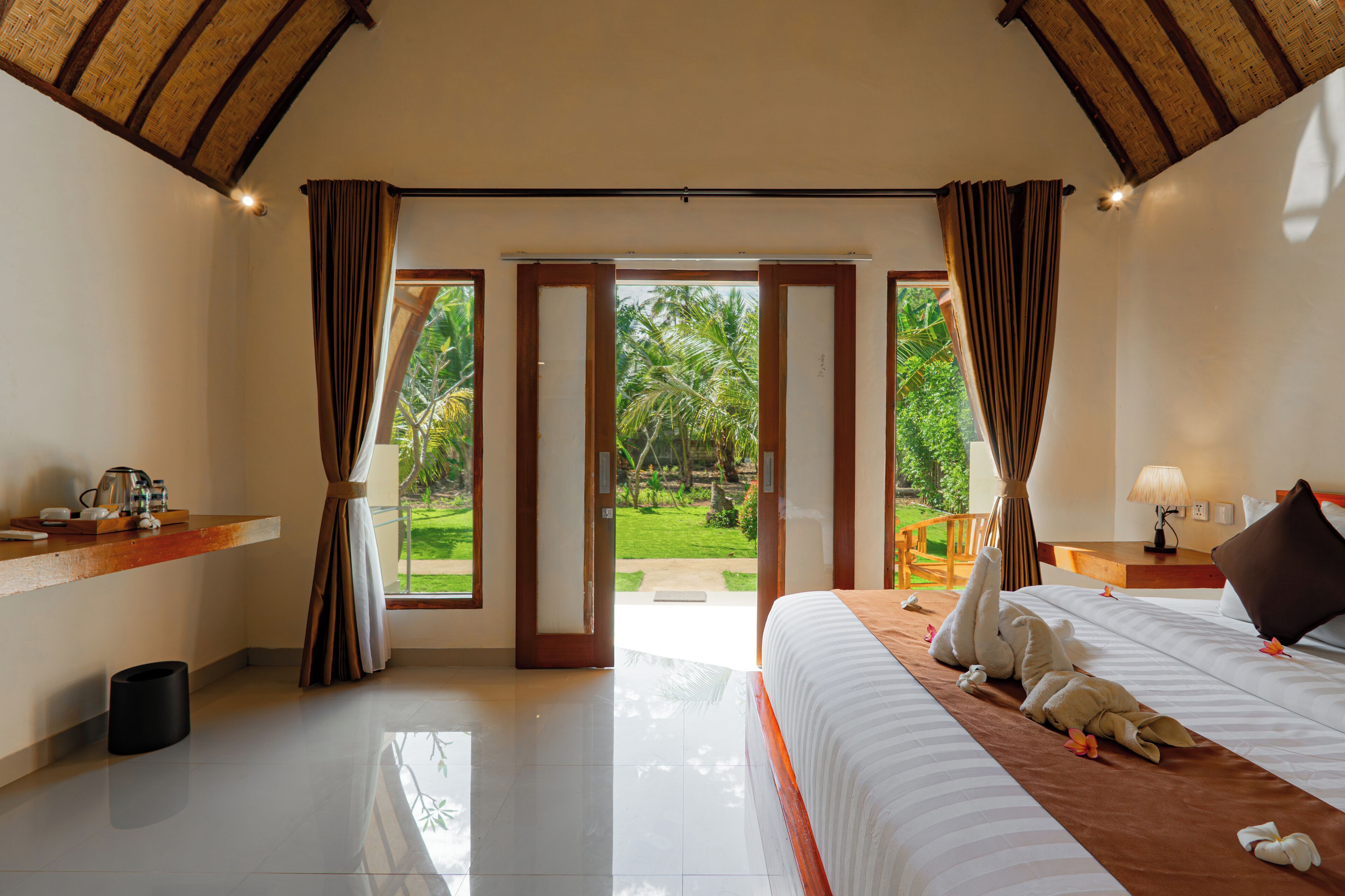 Una habitación de hotel con cama, mesita de noche, ventana con cortinas y vista a un jardín.