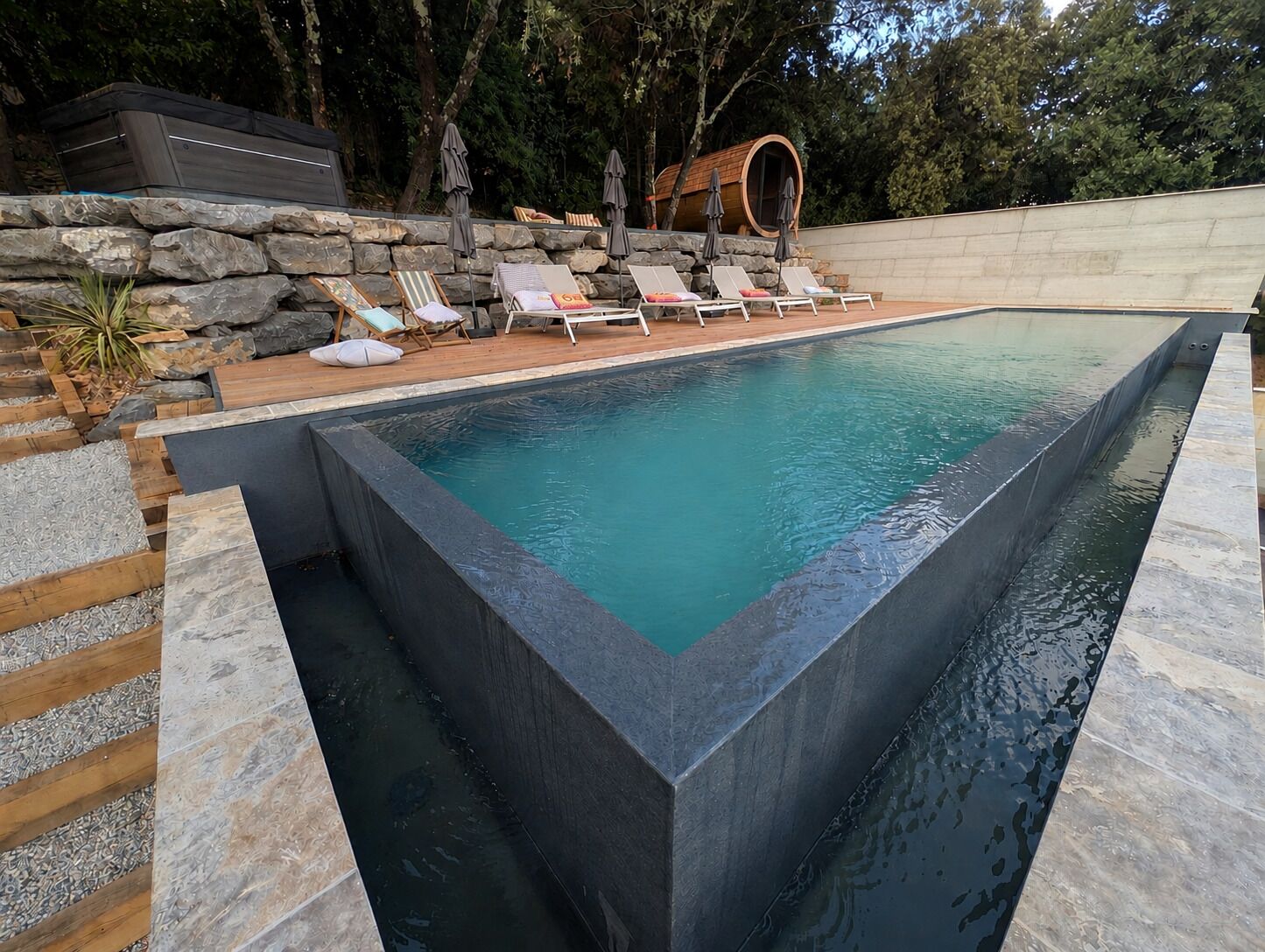 A rectangular swimming pool with a stone border, surrounded by lounge chairs and a stone wall.