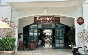 A entrada do JMO Hotel & Spa, com fachada de vidro transparente, uma placa sobre a entrada e área de estar ao ar livre.