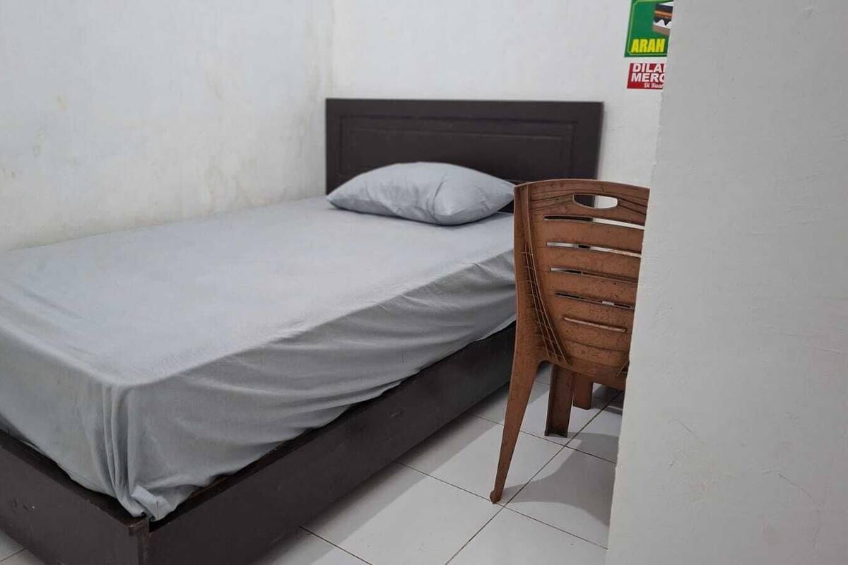 Een eenpersoonsbed met een grijze matras en een houten hoofdbord, een grijze kussen, en een houten stoel in een kamer met witte wanden en betegelde vloer.