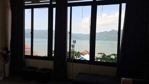 A room with a large window overlooking a body of water and mountains, with a bed and a small table. - Penginapan Remaja Morowali (Kolonodale)