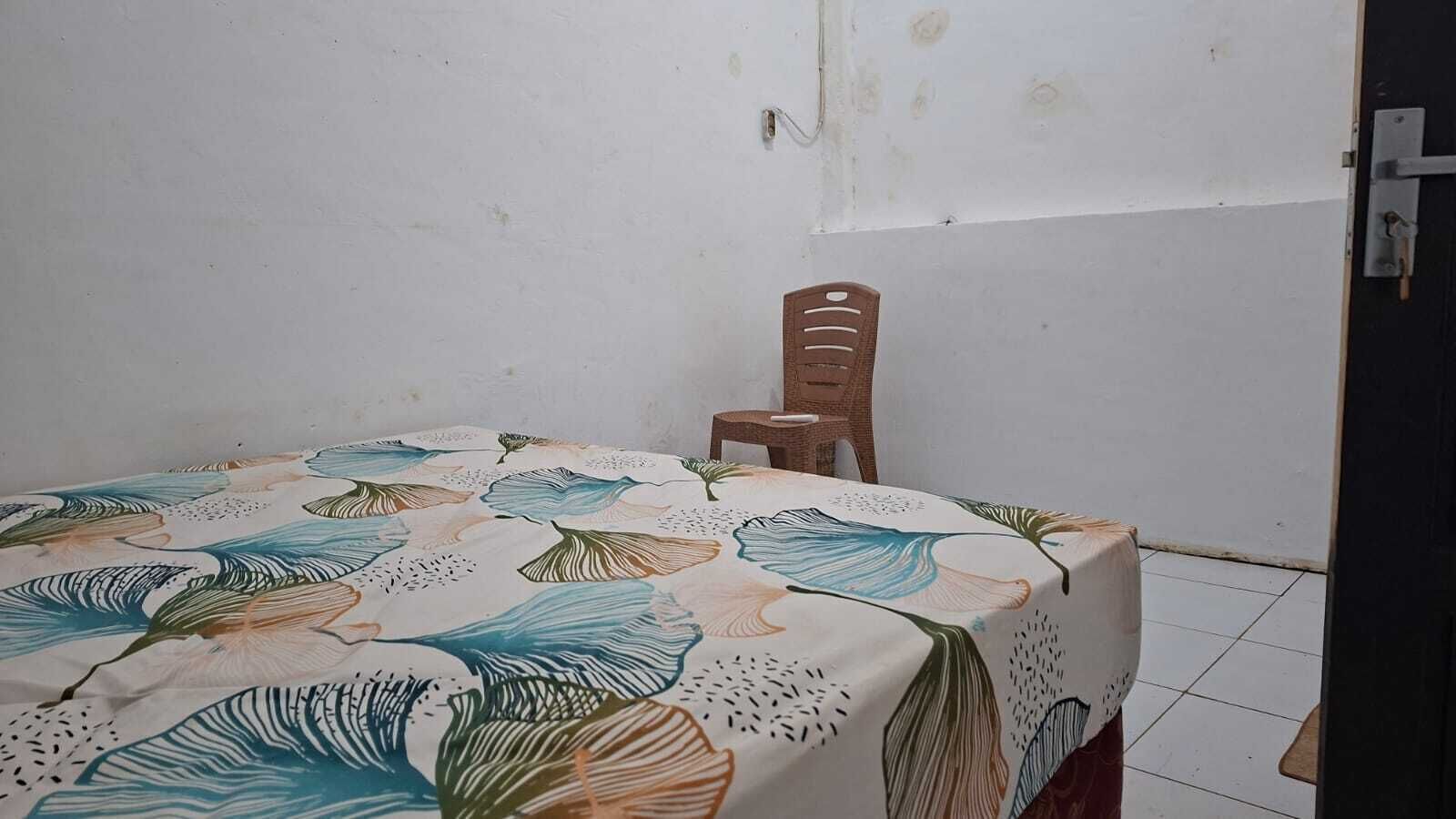 Una habitación pequeña con una cama cubierta por una colcha de estampado floral, una silla de madera y una puerta con manija metálica.