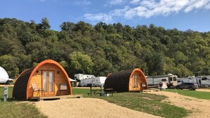 Dos cabañas de madera con techo arqueado y una mesa de picnic frente a ellas, ubicadas frente a una ladera boscosa.