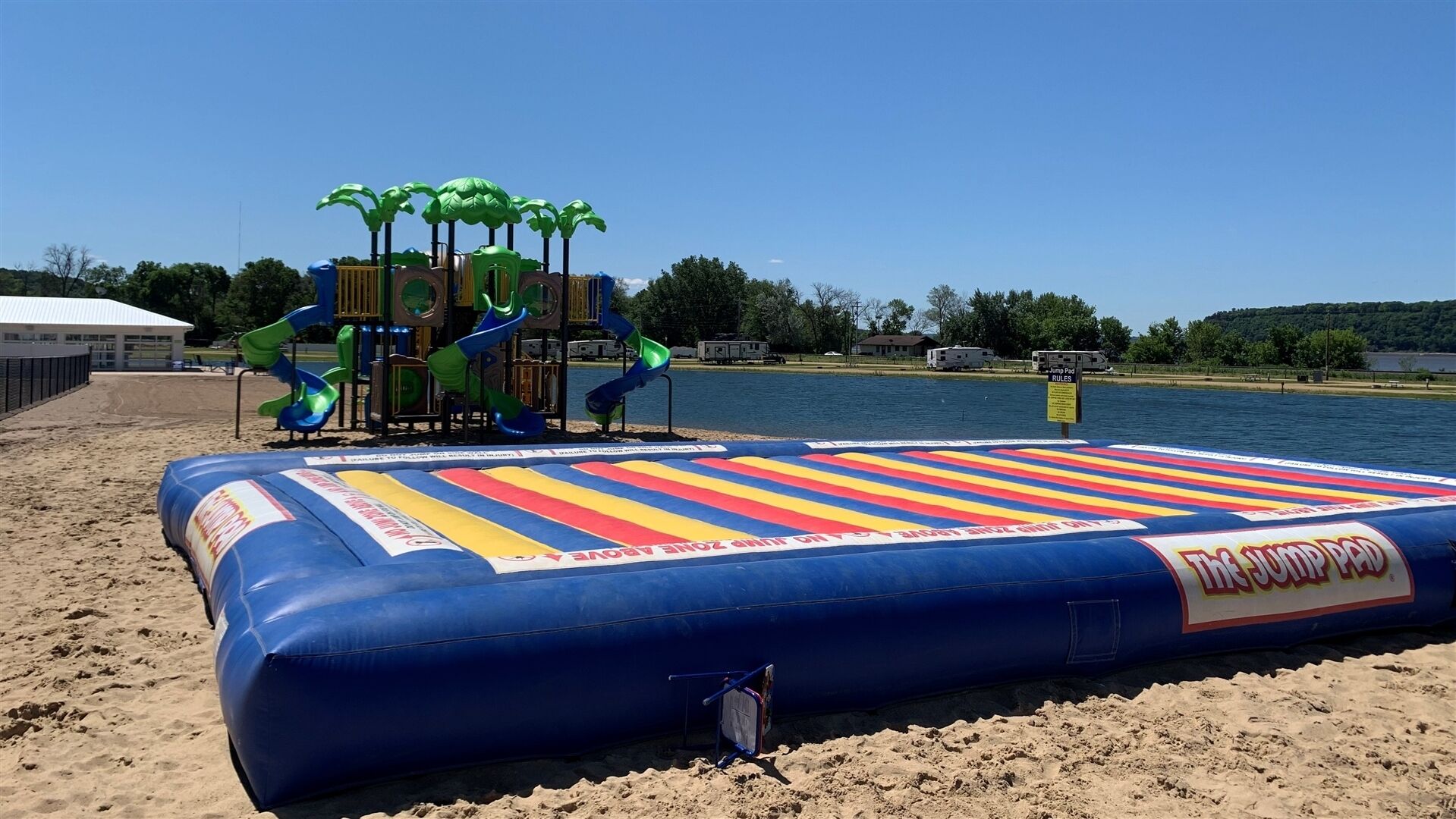 Une plage avec un grand tapis de saut gonflable, une aire de jeux avec des toboggans et une étendue d’eau en arrière-plan.
