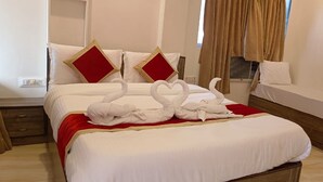 A hotel room with a bed, a nightstand, a bedside table, a window with curtains, and a small table with a folded towel. - Shagun Resort & Water Park (Igatpuri)