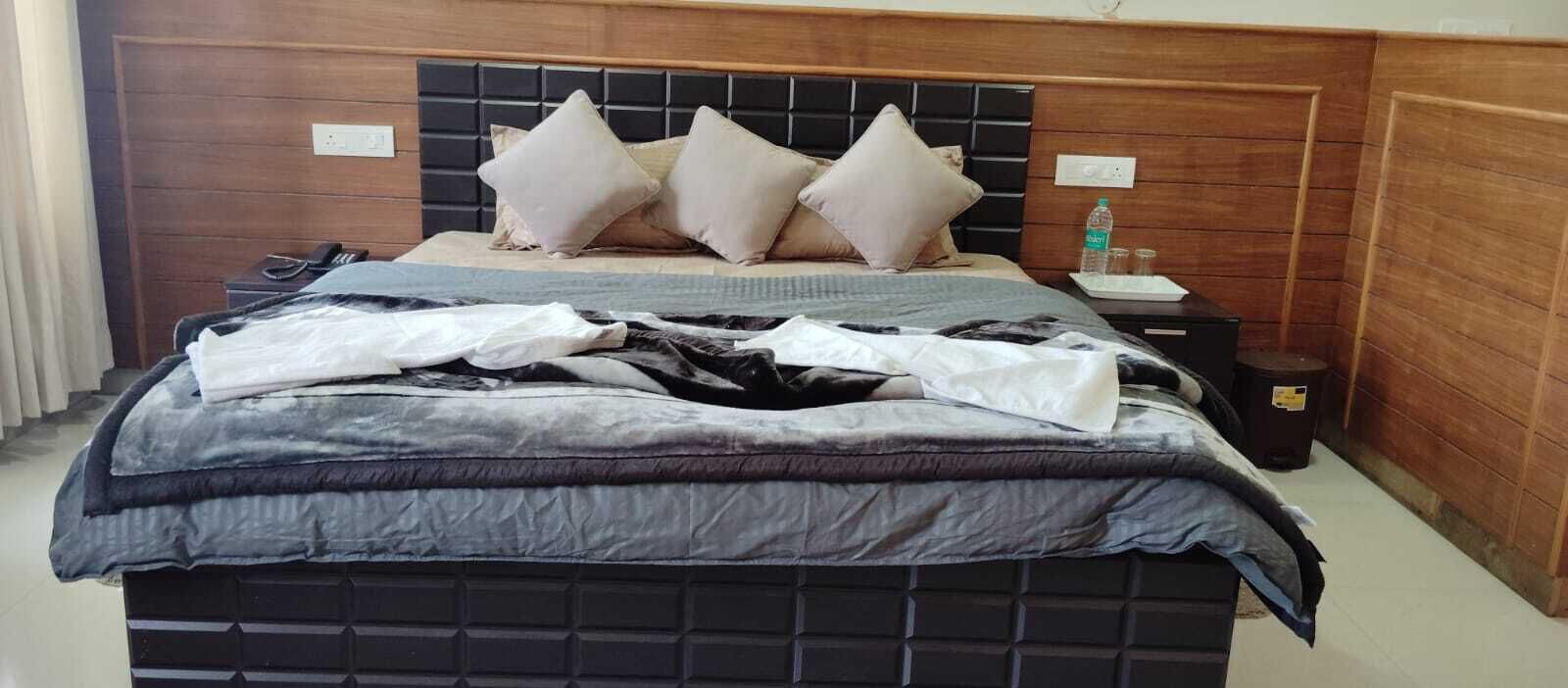 Ein ordentlich bezogenes Bett mit dunklem Kopfteil, ein Nachttisch mit Flasche und Gläsern sowie ein Papierkorb.