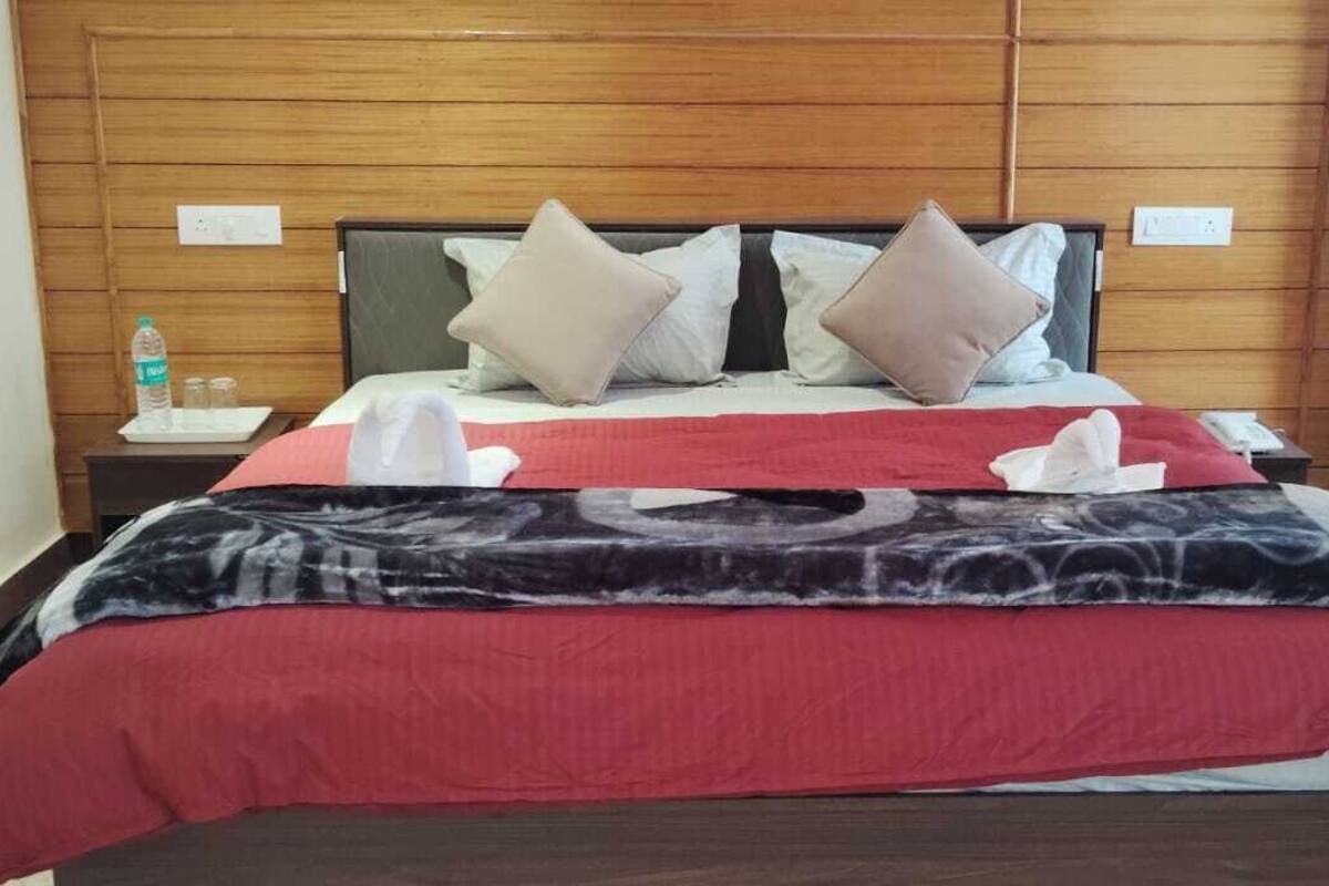 Habitación de hotel con cabecera de madera, cama con manta roja y negra, dos almohadas, mesita de noche con una botella y vasos, y un buró con un teléfono.