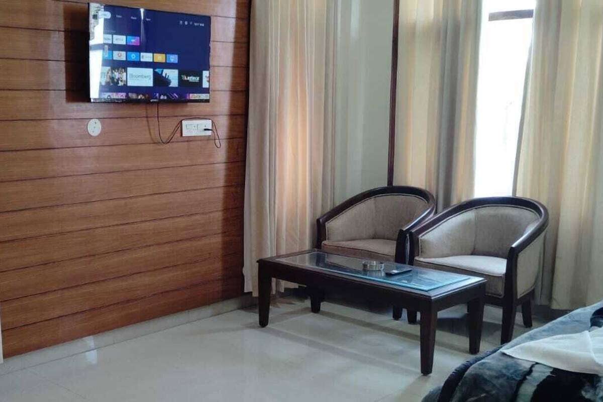 Habitación de hotel con pared de madera, un televisor de pantalla plana sobre una mesa de madera, dos sillas, una cama y una ventana con cortinas.