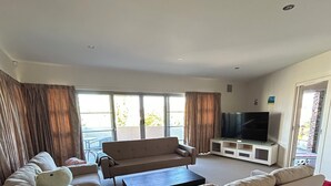 A living room with a curved sofa, a flat-screen TV, a coffee table, and a sliding glass door leading to a balcony. - House is near to university, AIS, mountain, lake and CBD. Family - friendly. (Bruce)