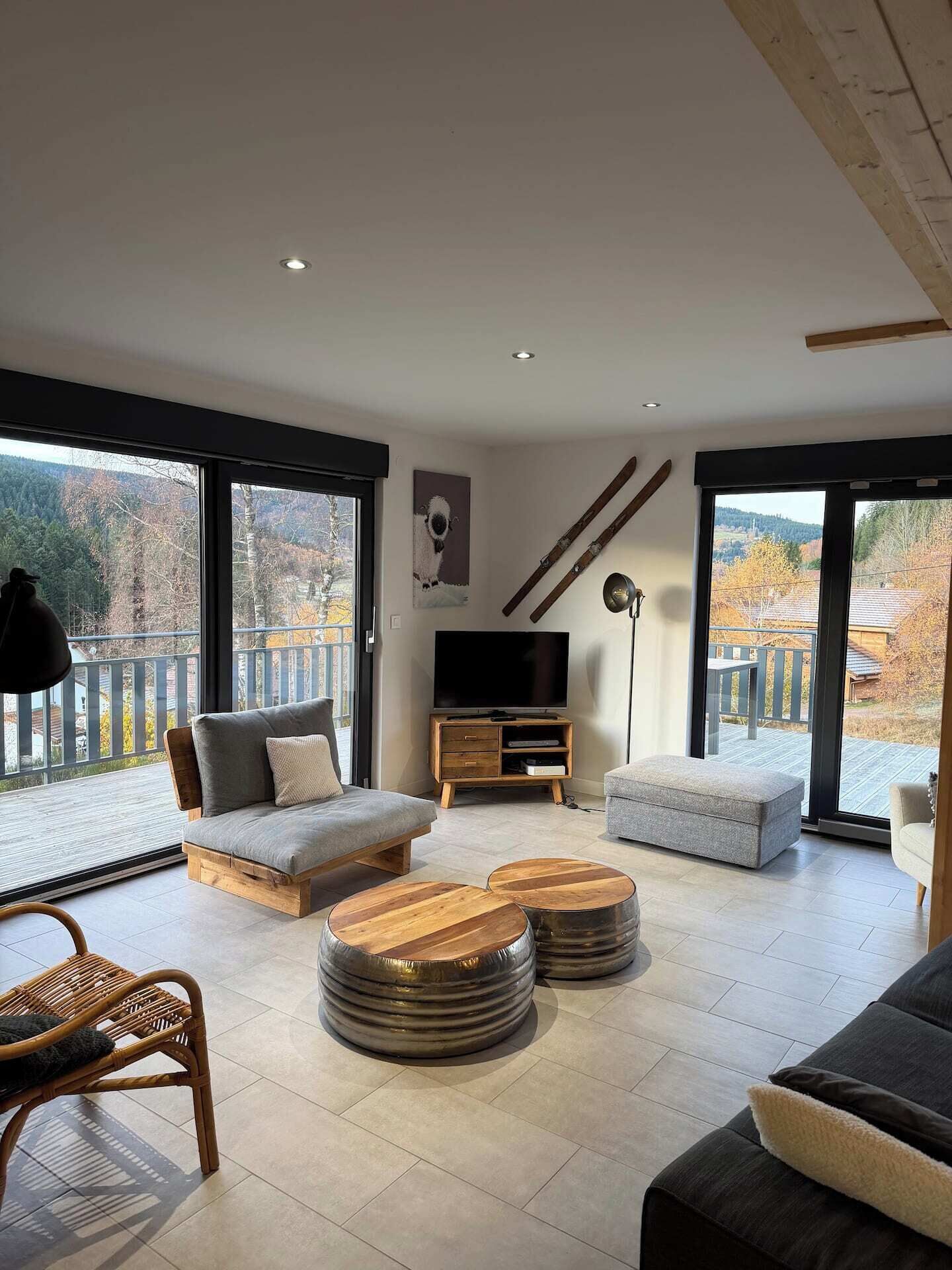 A modern living room with a sofa, ottoman, and coffee table, a TV, and large windows with a view of trees and a balcony.