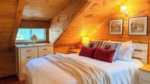 Un dormitorio acogedor con techo de madera, una cama con ropa blanca y almohadas rojas, una mesita de noche con lámpara y cuadros en la pared.