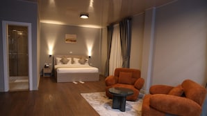 Une chambre d’hôtel comprenant un lit, un coin salon avec un fauteuil et une petite table, une table de chevet et une lampe fixée au mur.