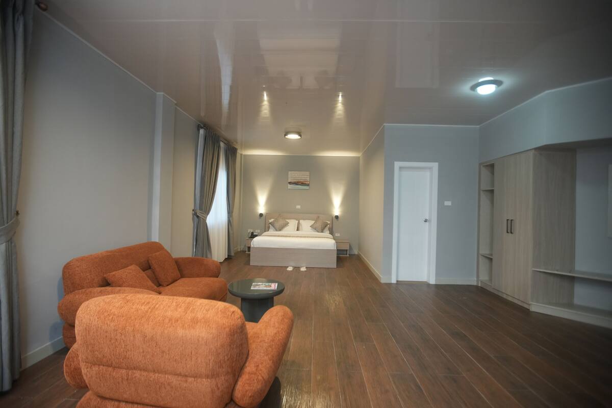 A modern hotel room with a large bed, a seating area with orange chairs, a small table, and a wardrobe.