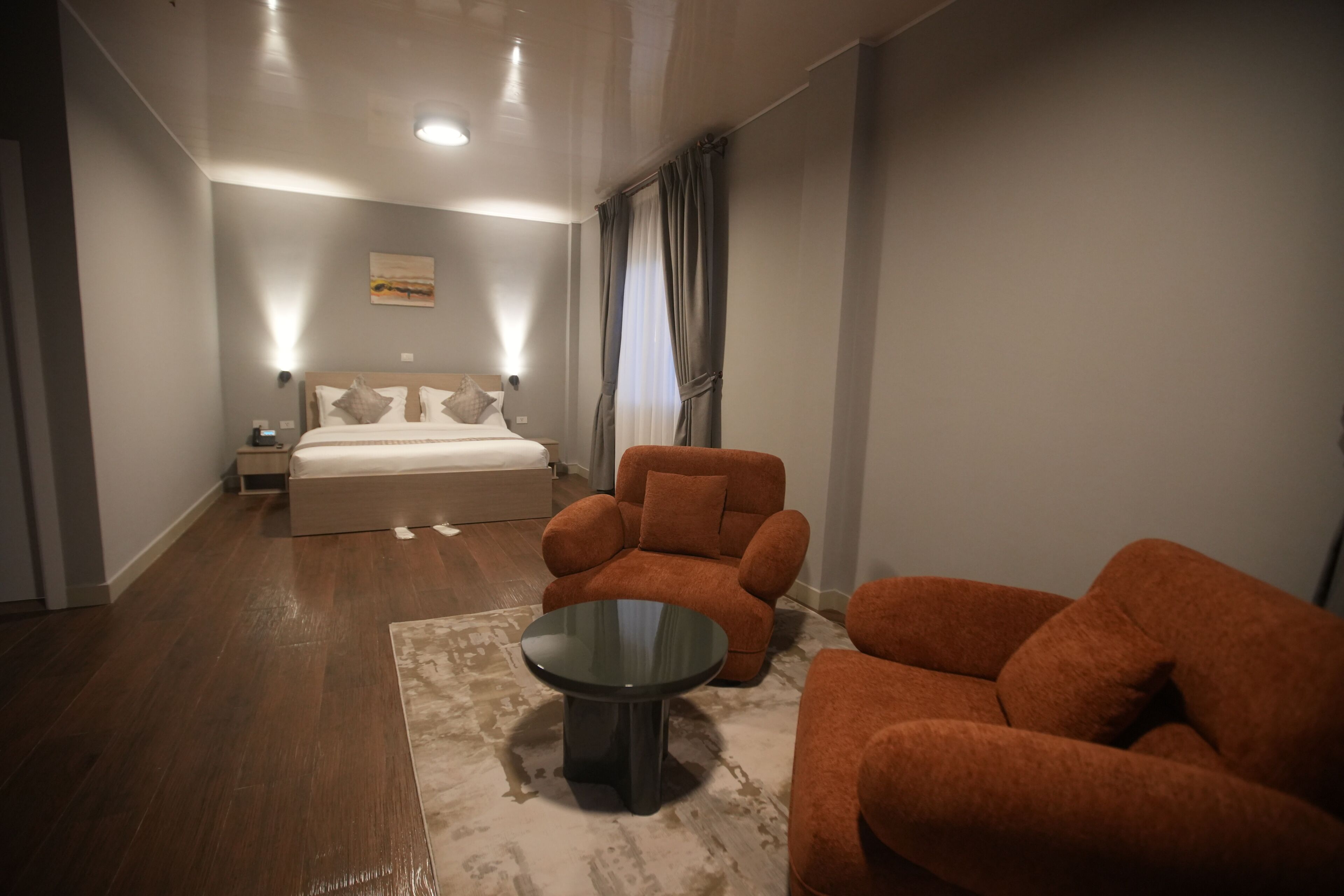 Habitación de hotel con cama, mesita de noche, mesa, dos sillones y una lámpara fijada a la pared.