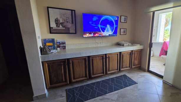 A kitchen with wooden cabinets, a tiled floor, a black rug, a television displaying a cityscape, and a door leading outside. - Cozy 1-bedroom condo in fantastic San José del Cabo with AC. Close to the beach (San José del Cabo)