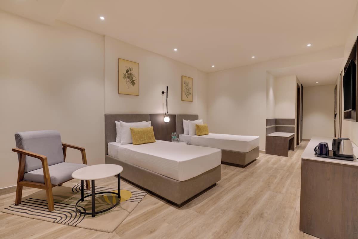 Habitación de hotel con dos camas, zona de estar con una silla y mesa, kitchenette y obras de arte en las paredes.