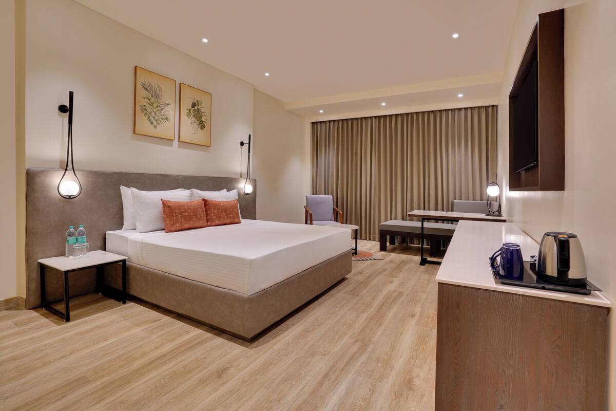 Una habitación de hotel con cama, mesitas de noche, escritorio, silla, televisión y un ventanal grande con cortinas.