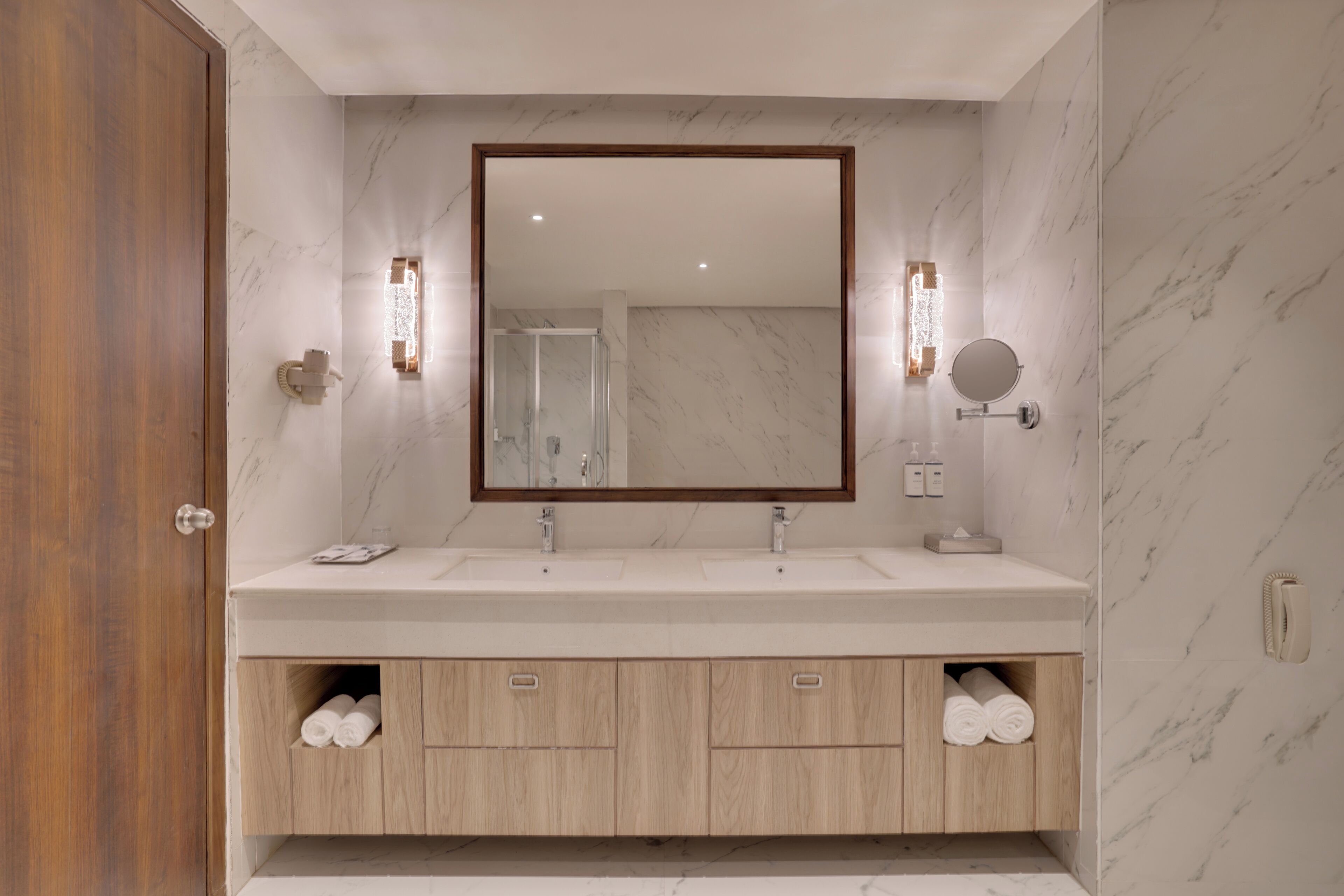 Une salle de bain avec une porte en bois, deux lavabos, un grand miroir et des luminaires fixés au mur.