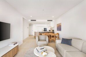 A modern living room with a flat-screen TV, a sofa, a coffee table, and a dining area in the background. - The Coastal Luxe Haven Coolangatta (Coolangatta)
