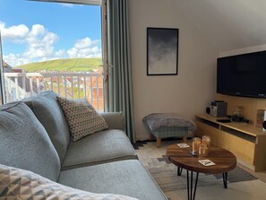 Un salon moderne avec un canapé, une petite table, un téléviseur et une vue sur un paysage verdoyant, visible depuis un balcon.