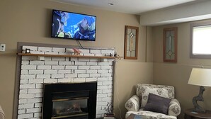 A living room with a fireplace, a television mounted above it, a patterned armchair, a small table with a lamp, and a rug on the floor.