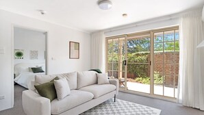 A modern living room with a sofa, a checkered rug, a sliding glass door leading to a garden, and a bed in the background. - Modern Neutral Haven with Premium Amenities (Griffith)