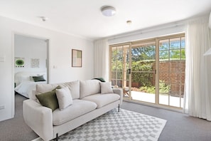 A modern living room with a sofa, a checkered rug, a sliding glass door leading to a garden, and a bed in the background. - Modern Neutral Haven with Premium Amenities (Griffith)